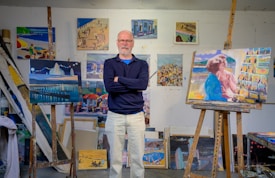 An older man stands confidently with arms crossed in an art studio. The walls are adorned with various colorful paintings displaying beach scenes and abstract art. Two finished canvases on easels feature a night landscape and a woman by the sea.
