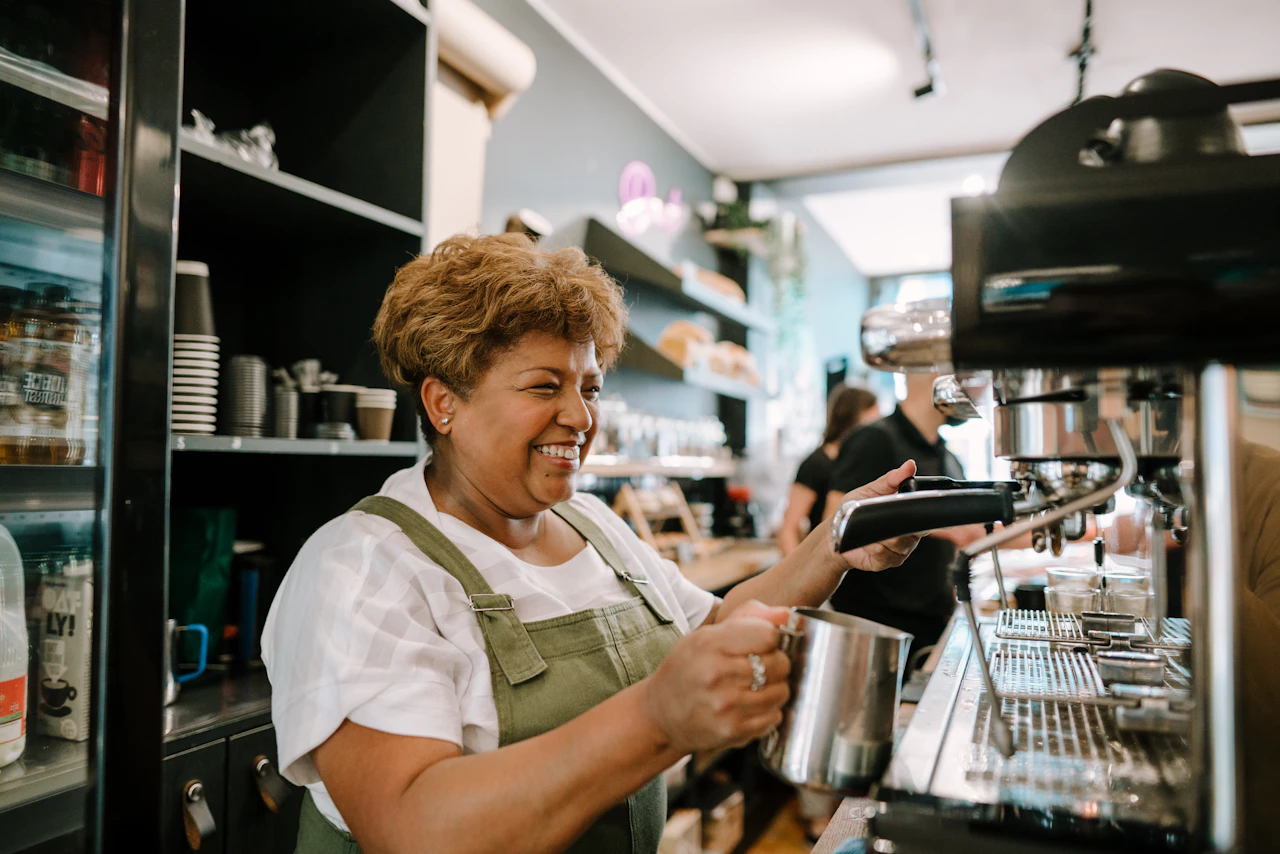 Smiling woman in a green apron steams milk at the espresso machine in a bright café.