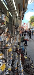 A bustling street market scene with various metal cookware and kettles displayed for sale on the left side of a shop. Price tags are visibly attached to many items. Several people are walking and browsing along the street. A seated man in the front appears to be taking a break. Red flags hang along the street, adding a touch of color to the lively atmosphere.