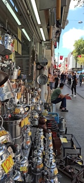 A bustling street market scene with various metal cookware and kettles displayed for sale on the left side of a shop. Price tags are visibly attached to many items. Several people are walking and browsing along the street. A seated man in the front appears to be taking a break. Red flags hang along the street, adding a touch of color to the lively atmosphere.