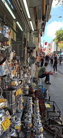 A bustling street market scene with various metal cookware and kettles displayed for sale on the left side of a shop. Price tags are visibly attached to many items. Several people are walking and browsing along the street. A seated man in the front appears to be taking a break. Red flags hang along the street, adding a touch of color to the lively atmosphere.