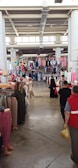 A bustling indoor market with various clothing items hanging from racks and displayed on tables. People are browsing, including some wearing traditional clothing. The ceiling is high with visible structural beams, and natural light filters through windows.