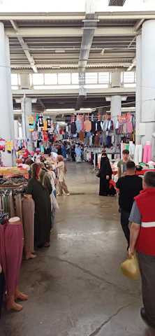 A bustling indoor market with various clothing items hanging from racks and displayed on tables. People are browsing, including some wearing traditional clothing. The ceiling is high with visible structural beams, and natural light filters through windows.