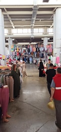 A bustling indoor market with various clothing items hanging from racks and displayed on tables. People are browsing, including some wearing traditional clothing. The ceiling is high with visible structural beams, and natural light filters through windows.