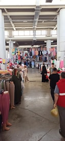 A bustling indoor market with various clothing items hanging from racks and displayed on tables. People are browsing, including some wearing traditional clothing. The ceiling is high with visible structural beams, and natural light filters through windows.