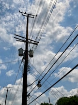 A wooden utility pole with multiple power lines stretches across a partly cloudy blue sky. Several transformers and a street lamp are attached to the pole.