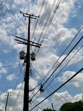 A wooden utility pole with multiple power lines stretches across a partly cloudy blue sky. Several transformers and a street lamp are attached to the pole.