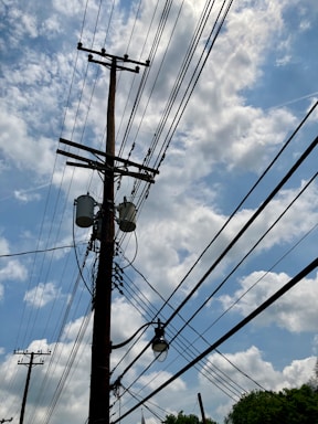 A wooden utility pole with multiple power lines stretches across a partly cloudy blue sky. Several transformers and a street lamp are attached to the pole.