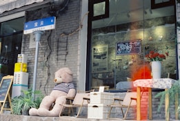 A large teddy bear wearing a striped shirt sits on a chair outside a shop. The shop has a brick exterior with a large glass window displaying various posters and flowers. There are also some chairs, a table with a floral tablecloth, and a few plants near the entrance. A street sign in blue, written in a non-English script, is visible above the teddy bear.