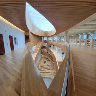 The image captures an interior space with a modern architectural design, featuring curved wooden structures and a high ceiling with a skylight. There are multiple floors visible, each with bookshelves and a wooden ramp or pathway that curves through the space. The openness and use of light wood create a bright and airy atmosphere.