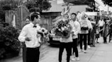 A group of people involved in what appears to be a ceremonial procession or event. Several individuals are holding intricately decorated flower arrangements, and there are people standing and observing around them. The setting seems to be an outdoor urban environment with buildings and a gate visible in the background.