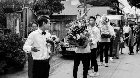 A group of people involved in what appears to be a ceremonial procession or event. Several individuals are holding intricately decorated flower arrangements, and there are people standing and observing around them. The setting seems to be an outdoor urban environment with buildings and a gate visible in the background.