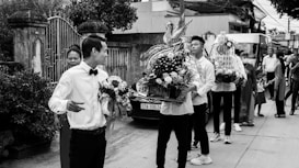 A group of people involved in what appears to be a ceremonial procession or event. Several individuals are holding intricately decorated flower arrangements, and there are people standing and observing around them. The setting seems to be an outdoor urban environment with buildings and a gate visible in the background.