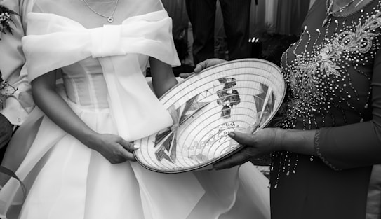 A woman in a detailed, embroidered dress is holding a traditionally decorated woven hat with intricate designs. Another woman, dressed in a formal gown with a large bow, stands beside her. The scene appears ceremonial.