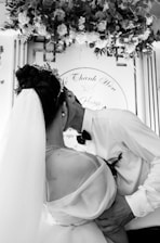 A couple embracing in a romantic gesture, with the bride wearing a veil and wedding dress, while the groom is in a shirt and bow tie. The background features floral decorations and scripted text.