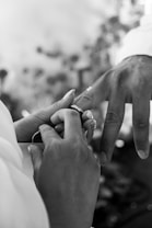 A close-up, black-and-white image depicts a person placing a ring on another person's finger. The focus is on the hands, which are gently engaged in the exchange. The background is softly blurred, emphasizing the intimacy of the moment.