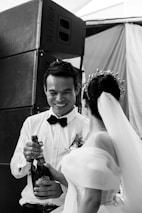 A couple is dressed in formal attire, with one person in a wedding dress holding a champagne bottle while the other person, wearing a bow tie and boutonniere, smiles. They stand in front of large speakers in what appears to be a festive setting.