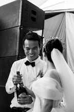 A couple is dressed in formal attire, with one person in a wedding dress holding a champagne bottle while the other person, wearing a bow tie and boutonniere, smiles. They stand in front of large speakers in what appears to be a festive setting.