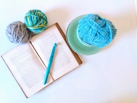An open book lies on a light surface surrounded by two skeins of yarn in shades of blue, yellow, and green. A teal crochet hook rests on the book, and to the side is a third blue ball of yarn placed on a green plate.