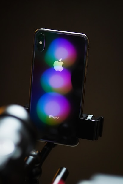 A smartphone with a prominent Apple logo on the back is mounted on a tripod. The device is reflecting vibrant circular light patterns in purple, blue, and green hues. The setting has a dark background that enhances the vivid colors.