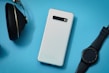 A white smartphone is placed on a light blue background, accompanied by a black smartwatch and a pair of black headphones. The smartphone is sleek with a modern design, and the smartwatch has a round face with a black strap, while the headphones have an over-ear design.