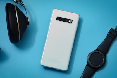 A white smartphone is placed on a light blue background, accompanied by a black smartwatch and a pair of black headphones. The smartphone is sleek with a modern design, and the smartwatch has a round face with a black strap, while the headphones have an over-ear design.