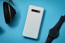 A white smartphone is placed on a light blue background, accompanied by a black smartwatch and a pair of black headphones. The smartphone is sleek with a modern design, and the smartwatch has a round face with a black strap, while the headphones have an over-ear design.