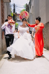 A joyful wedding scene depicts a bride in a white dress walking down a narrow alley, accompanied by a man holding a bouquet and a woman adjusting her large, colorful hat. The alley is framed by high walls, and several people can be seen in the background, smiling and celebrating.