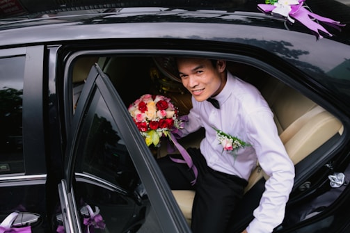 A man dressed in formal attire, including a white shirt and black bow tie, is seated inside a car. He holds a vibrant bouquet of flowers, predominantly composed of red and white roses with green foliage and purple ribbons. The car's interior features cream-colored seats, and the vehicle exterior is decorated with purple floral arrangements.
