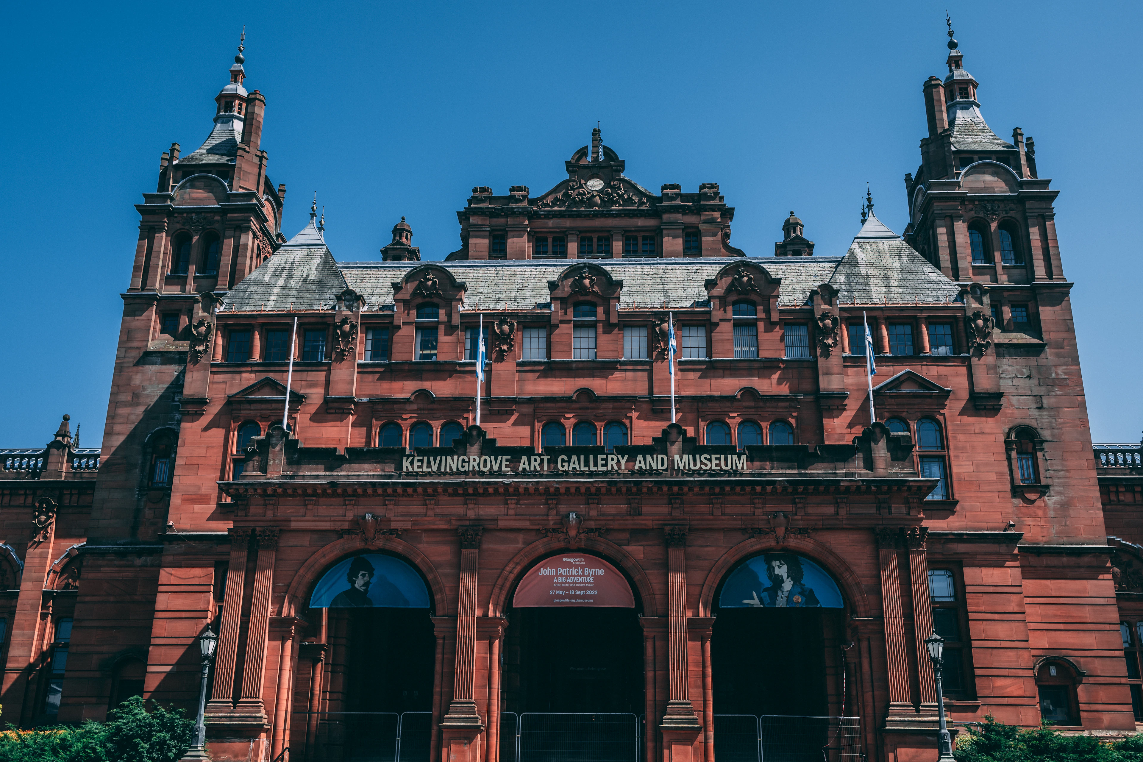 The red sandstone facade of Kelvingrove Art Gallery and Museum in Glasgow, its towers and carved stonework rising symmetrically against a deep blue summer sky, with the museum's name cut into the stone above the arched entrances.
