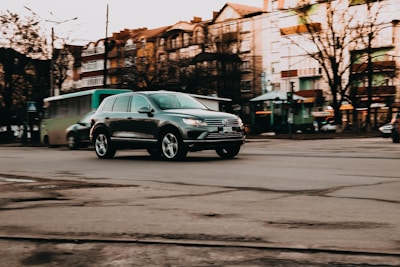 A modern SUV is driving through an urban street with old-style apartment buildings in the background. The road seems to have some cracks and rough patches. There's a bus in the background, and the overall setting suggests early evening with an overcast sky.