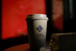 A branded coffee cup with a logo is placed on a surface. The background is predominantly red with blurred elements, providing a contrast to the cup. The lighting creates shadows and highlights the details on the cup, suggesting a warm ambiance.