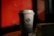 A branded coffee cup with a logo is placed on a surface. The background is predominantly red with blurred elements, providing a contrast to the cup. The lighting creates shadows and highlights the details on the cup, suggesting a warm ambiance.