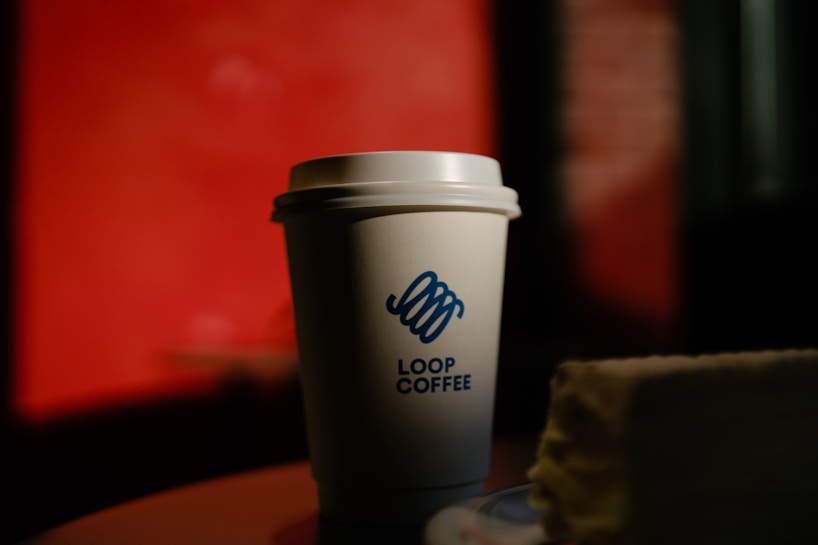 A branded coffee cup with a logo is placed on a surface. The background is predominantly red with blurred elements, providing a contrast to the cup. The lighting creates shadows and highlights the details on the cup, suggesting a warm ambiance.