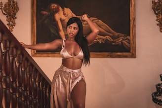 A woman stands gracefully near a staircase, wearing a silky outfit. Behind her, there is a classical painting featuring a reclining figure. Ornate wall decorations and wooden staircase rails add to the sophisticated atmosphere.