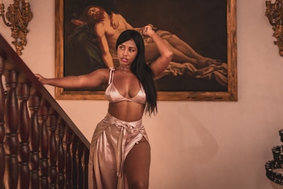 A woman stands gracefully near a staircase, wearing a silky outfit. Behind her, there is a classical painting featuring a reclining figure. Ornate wall decorations and wooden staircase rails add to the sophisticated atmosphere.