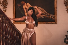 A woman stands gracefully near a staircase, wearing a silky outfit. Behind her, there is a classical painting featuring a reclining figure. Ornate wall decorations and wooden staircase rails add to the sophisticated atmosphere.
