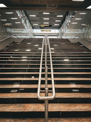 A wide staircase with polished stone steps and a central metal railing, leading upwards to a platform with glass walls and signs. Overhead, fluorescent lights are embedded in a patterned ceiling.