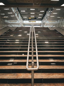 A wide staircase with polished stone steps and a central metal railing, leading upwards to a platform with glass walls and signs. Overhead, fluorescent lights are embedded in a patterned ceiling.