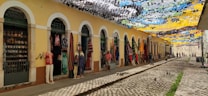An outdoor market scene with vibrant decorative banners overhead casting colorful shadows. The market stalls are housed in a yellow building with archways. Various clothing items and textiles are displayed outside the shops, along with decorative items. The cobblestone street adds a rustic charm to the setting.