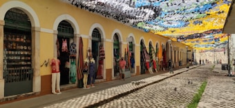 An outdoor market scene with vibrant decorative banners overhead casting colorful shadows. The market stalls are housed in a yellow building with archways. Various clothing items and textiles are displayed outside the shops, along with decorative items. The cobblestone street adds a rustic charm to the setting.