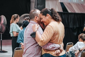 A group of people are socializing indoors. A man and a woman are embracing and appear to be engaged in a heartfelt conversation. Other people in the background are also interacting with one another. The atmosphere is casual and warm.