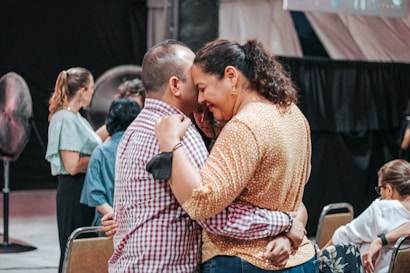 A group of people are socializing indoors. A man and a woman are embracing and appear to be engaged in a heartfelt conversation. Other people in the background are also interacting with one another. The atmosphere is casual and warm.