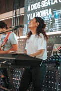 A woman with curly hair is joyfully playing a keyboard on stage. She is wearing a white t-shirt and glasses, with a microphone in front of her. Behind her is a screen displaying text in Spanish and lights creating a lively background. Another person is playing a guitar beside her.