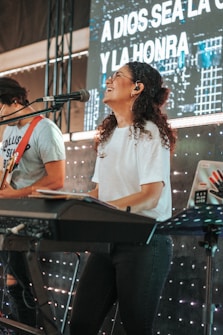 A woman with curly hair is joyfully playing a keyboard on stage. She is wearing a white t-shirt and glasses, with a microphone in front of her. Behind her is a screen displaying text in Spanish and lights creating a lively background. Another person is playing a guitar beside her.
