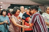 A group of people is gathered in what appears to be a social event or meeting. One person is holding a microphone and shaking hands with another individual, suggesting an introduction, speech, or presentation taking place. The expressions vary from attentive to cheerful. People in the background seem engaged, adding to the lively atmosphere.