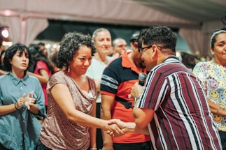 A group of people is gathered in what appears to be a social event or meeting. One person is holding a microphone and shaking hands with another individual, suggesting an introduction, speech, or presentation taking place. The expressions vary from attentive to cheerful. People in the background seem engaged, adding to the lively atmosphere.