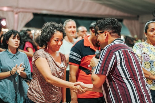 A group of people is gathered in what appears to be a social event or meeting. One person is holding a microphone and shaking hands with another individual, suggesting an introduction, speech, or presentation taking place. The expressions vary from attentive to cheerful. People in the background seem engaged, adding to the lively atmosphere.