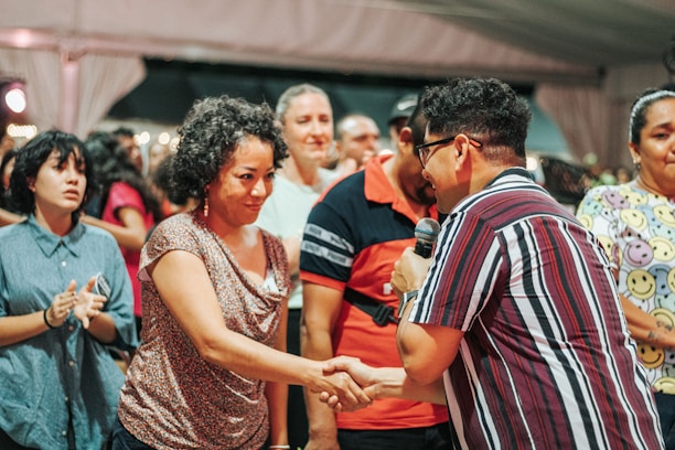 A group of people is gathered in what appears to be a social event or meeting. One person is holding a microphone and shaking hands with another individual, suggesting an introduction, speech, or presentation taking place. The expressions vary from attentive to cheerful. People in the background seem engaged, adding to the lively atmosphere.
