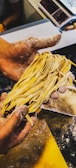 A person with flour-dusted hands is holding a bunch of freshly made pasta. The background shows a countertop sprinkled with flour and various kitchen tools.